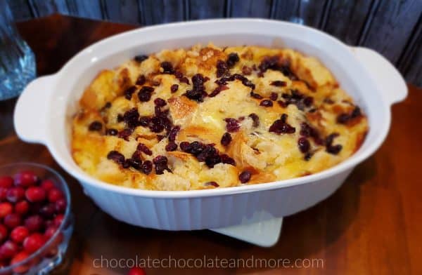 A large white dish of cranberry brie bread pudding on a hot plate next to a bowl of cranberries.