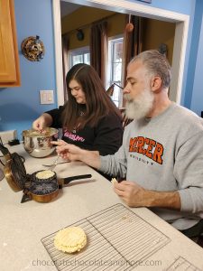 Father and daughter sitting at a kitchen counter make pizzelles on a pizzelle iron.