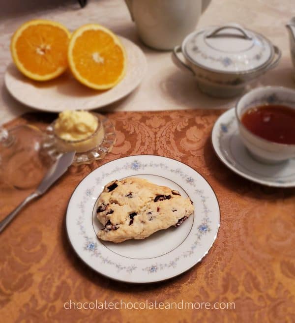 A place setting of china with a cranberry scone with orange butter on the side. There is also a cup of tea and a sliced orange.