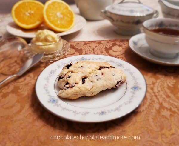 A place setting of china with a cranberry scone with orange butter on the side. There is also a cup of tea and a sliced orange.
