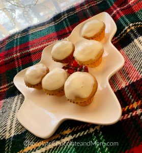 6 mini Eggnog Cream Cheese Cupcakes sitting on a green, red, and white blanket with a white lace curtain in the background.