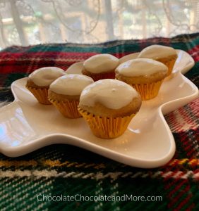 6 mini Eggnog Cream Cheese Cupcakes sitting on a green, red, and white blanket with a white lace curtain in the background.