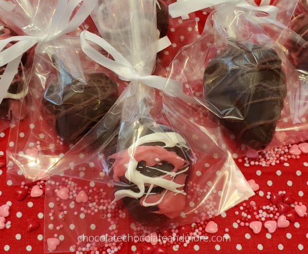 chocolate covered strawberries decorated with pink and white chocolate wrapped in cellophane bags tied with ribbons on a red background covered with candy hearts