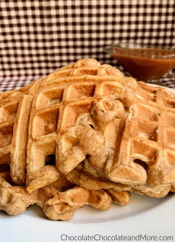Cinnamon Applesauce Waffles sitting on a white circular plate and a clear gravy boat with cinnamon applesauce glaze sitting behind and a white and black checkered background.