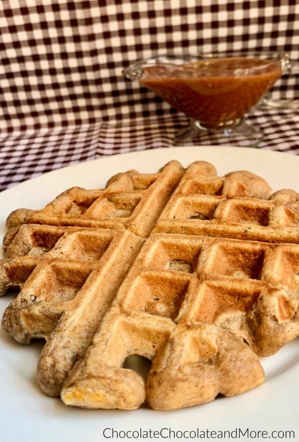 Cinnamon Applesauce Waffles sitting on a white circular plate and a clear gravy boat with cinnamon applesauce glaze sitting behind and a white and black checkered background.