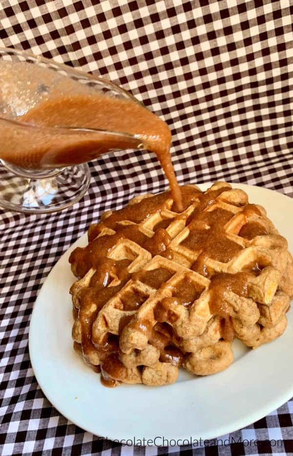 Cinnamon Applesauce Waffles sitting on a white circular plate and a clear gravy boat with cinnamon applesauce glaze being poured on top and a white and black checkered background.