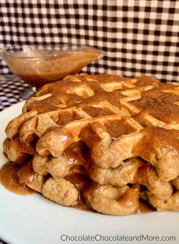 Cinnamon Applesauce Waffles sitting on a white circular plate and a clear gravy boat with cinnamon applesauce glaze sitting behind and a white and black checkered background.