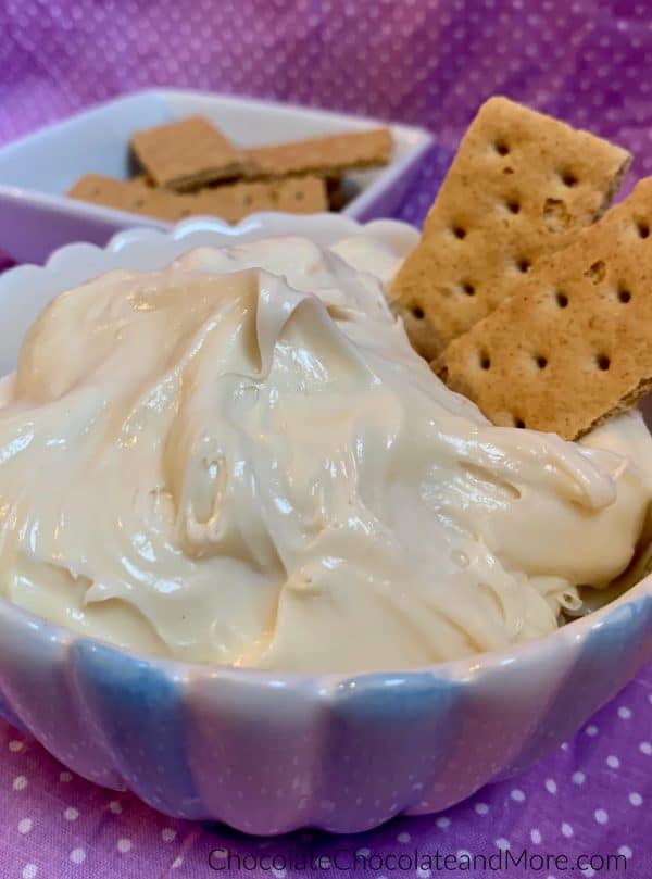 A bowl of 3-Ingredient Cheesecake Dip with 2 graham crackers sticking out and a bowl of graham crackers behind it with a purple and white polka dot background.