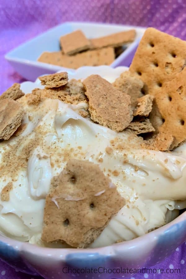 A bowl of 3-Ingredient Cheesecake Dip topped with crushed graham crackers and a bowl of graham cracker behind it with a purple and white polka dot background.