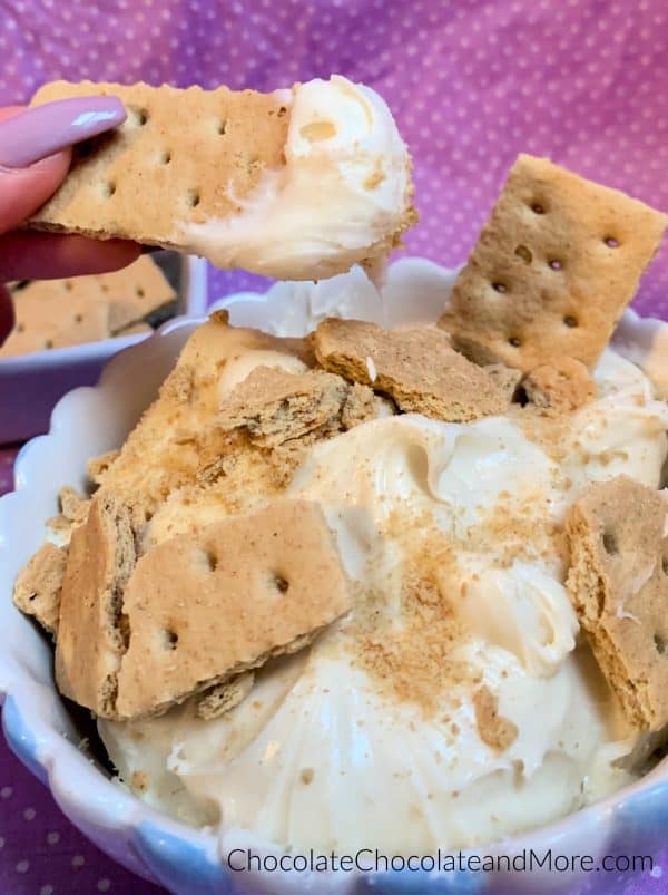 A bowl of 3-Ingredient Cheesecake Dip topped with crushed graham crackers with a graham cracker scooping some out with a purple and white polka dot background.