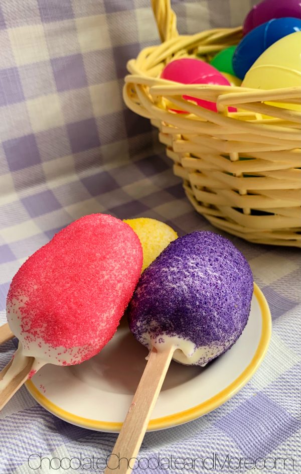 3 Easter Egg Cake Pops (one with yellow sprinkles, one with pink, and one with purple) sitting on a white with yellow rimmed plate and a yellow basket filled with plastic colored easter eggs sitting on a purple and white plaid background.