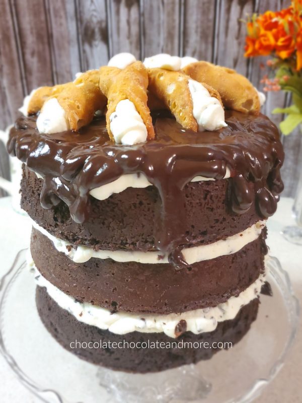 a very tall triple layer chocolate cake dripping with chocolate fudge and topped with cannolis