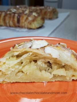 A slice of almond puff pastry on an orange plate in front of two loaves