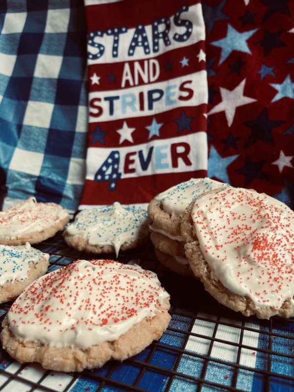 8 Cream Cheese Frosted Sugar Cookies sitting on a cooling rack on a checkered blue and white background with a "stars and stripes for ever" towel in the back.