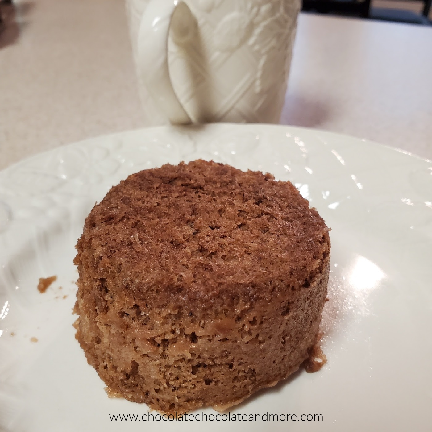a small round chocolate cake on a plate with a mug in the background