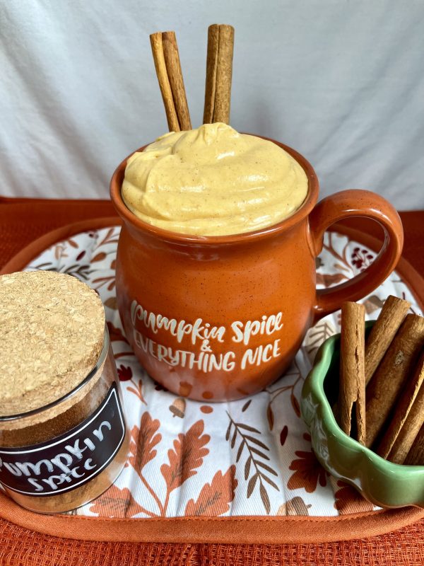 Pumpkin Spice Cheesecake Dip in an orange mug with a jar of pumpkin spice and a bowl of cinnamon sticks in front of it.