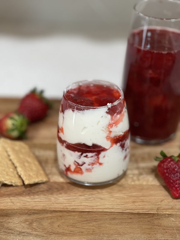 Strawberry Shortcake DIp in a cup sitting on a wooden cutting board with graham crackers and strawberries lying next to it and strawberry syrup in a taller cup behind.