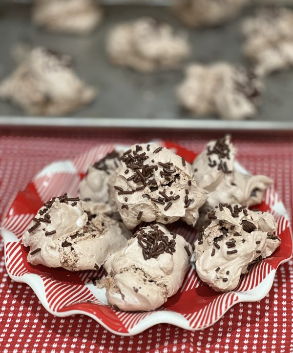 Hot Cocoa Meringue Cookies in a red plaid bowl in front of a cookie sheet with the rest of the cookies on it