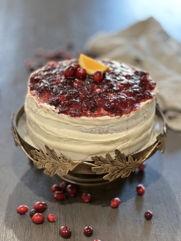 Cranberry Orange Triple Layer Cake on a cake stand with cranberrys on the table.