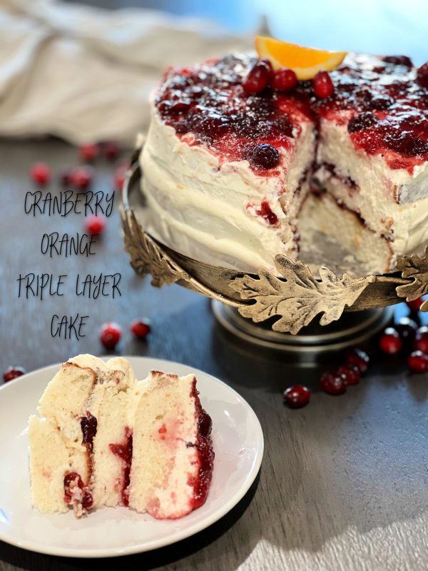 Cranberry Orange Triple Layer Cake with a piece cut out and palced on a white plate with cranberrys on the table